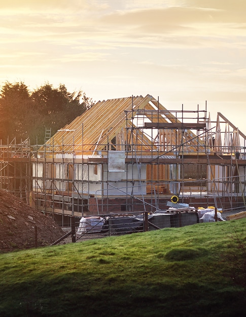 Maison en construction au lever du soleil, avec charpente en bois et échafaudages. Pelouse verte au premier plan.