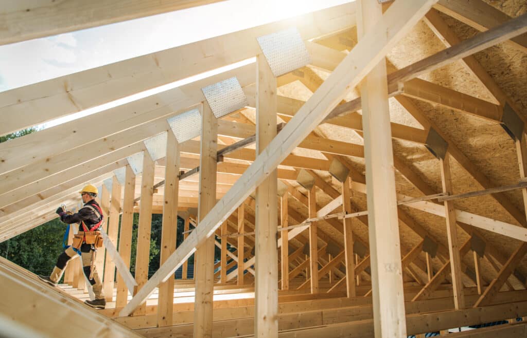 Charpentier avec casque et harnais travaillant sur la charpente en bois d'une maison en construction, sous le soleil.