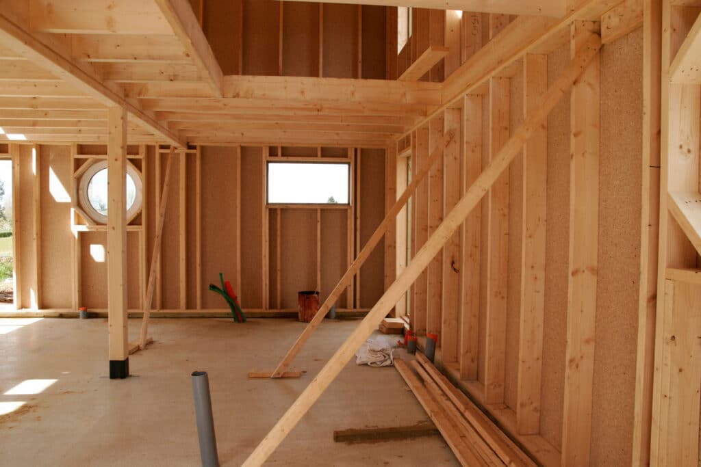 Intérieur d'une maison en construction, avec l'ossature bois et les panneaux OSB apparents. Fenêtres ronde et rectangulaire, sol en béton.