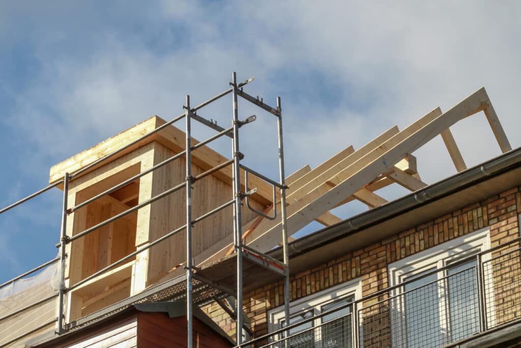 Construction de lucarne et charpente en bois sur un toit d'immeuble, avec échafaudage, sous un ciel bleu nuageux.