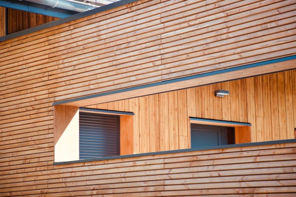 Façade moderne en bois clair à lames horizontales, avec une grande ouverture révélant des fenêtres et un volet roulant. Architecture.