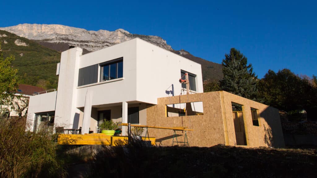 Maison moderne blanche en construction avec une extension en bois, sous un ciel bleu. Une grande montagne rocheuse domine l'arrière-plan.