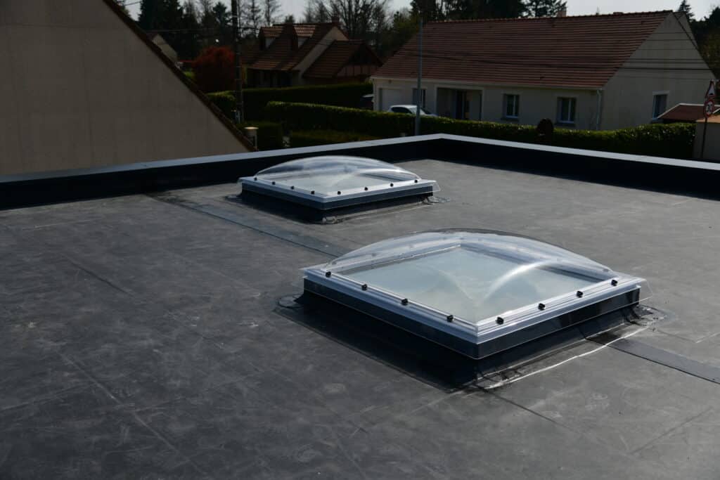 Toit plat étanche noir avec deux puits de lumière Velux installés. En arrière-plan, maisons résidentielles et panneau 30 km/h.